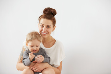 Young attractive mother with her one years old little son dressed in pajamas.  Boy eating a fruit smoothie himself in the bedroom at the weekend together, warm and cozy scene. Selective focus.