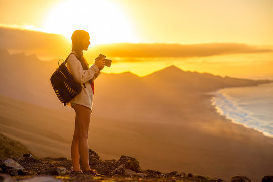 Young female traveler with backpack and photo camera photographing beautiful Cofete coastline with mountains on the sunset on Fuerteventura island in Spain. 