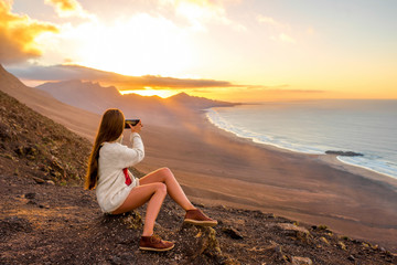 Fototapeta na wymiar Young woman photographing with smart phone beautiful landscape with beach and mountains on the sunset on Fuerteventura island in Spain