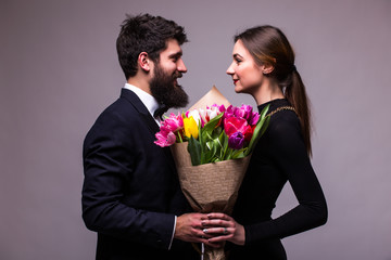 Portrait of young couple in love with bouquet of spring tulips posing dressed in classic clothes on white backround. Man with beard in Suit, Woman in black Dress look each other. 