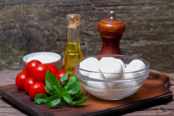 Italian cuisine. Mediterranean cuisine.  Traditional italian mozzarella cheese in glass bowl with basil, tomatoes and olive oil - caprese salad ingredients, on rustic wooden background. Rustic style