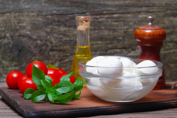 Italian cuisine. Mediterranean cuisine.  Traditional italian mozzarella cheese in glass bowl with basil, tomatoes and olive oil - caprese salad ingredients, on rustic wooden background. Rustic style