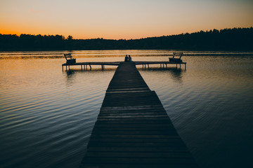 Obraz premium mężczyzna i kobieta siedzi o zachodzie słońca na drewnianym nabrzeżu nad jeziorem w lesie