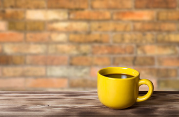 Coffee in yellow cup on wooden table opposite a defocused brick