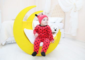 Маленький ребенок, одетый в красный костюм с черным в горошек. Детский интерьер. Белая светлая комната с кроваткой, луна игрушка, плюшевый мишка.