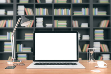 Blank laptop screen with cell phone on wooden table at bookcase