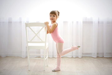 Little cute girl in pink leotard making new ballet movement near chair at dance studio