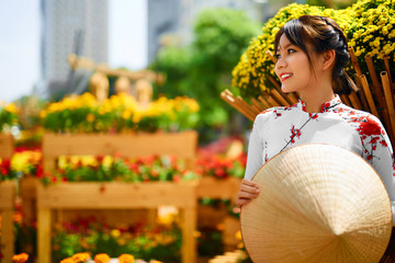 Culture Of Asia. Beautiful Happy Smiling Young Asian Woman Wearing Traditional White Ao Dai Dress ( Clothing ), Vietnamese Conical Hat ( Non La ) Leaf Hat In Flower Garden In Ho Chi Minh City, Vietnam - obrazy, fototapety, plakaty