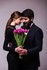 Portrait of young couple in love with bouquet of lila tulips posing dressed in classic clothes on grey backround. Man with beard in Suit, Woman in black Dress. The concept of Spring holiday