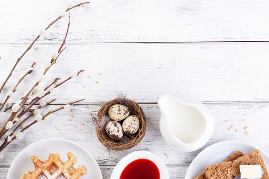 Easter breakfast with quail eggs, waffles, fruit jam, milk and sandwiches, with willow branch on a white wooden background, top view with copy space..