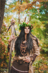 beautiful brunette with painted face, in a flower wreath and a wooden staff in his hand, looking...