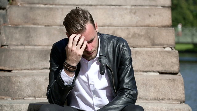 Trendy looking man sitting alone outside on steps alongside a river, wearing a black leather jacket and a white shirt. Looking away from camera with a sad look.