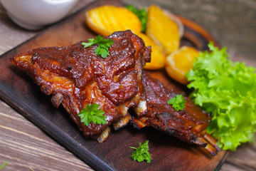 Appetizing rosy and barbecued lamb ribs seasoned with a barbecue sauce and served with fresh herbs and potatoes on an old rustic wooden chopping board