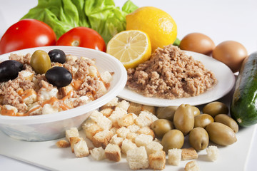 Takeaway tuna salad served in a white plastic bowl next to a white plate with tuna chunks, lemon, olives, croutons, and fresh eggs, cucumbers, tomatoes and lettuce.