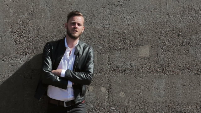 A man in his 20s wearing a black leather jacket and white shirt, leaning back against a wall of stone on a sunny summer day,