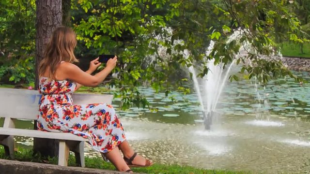 Blond Girl Sits on Bench Takes Photo of Fountain with Iphone