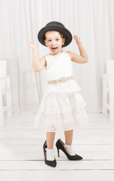 Adorable cute caucasian girl wearing white dress, black hat and black high heel shoes. Little girl trying to walk with big high heel shoes. Little girl fashionista in her mother's big heeled shoes