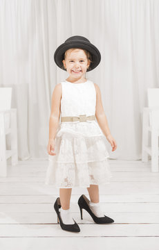 Adorable cute caucasian girl wearing white dress, black hat and black high heel shoes. Little girl trying to walk with big high heel shoes. Little girl fashionista in her mother's big heeled shoes