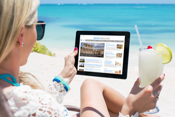 Woman reading news on tablet while relaxing on the beach. All website contents are made up.