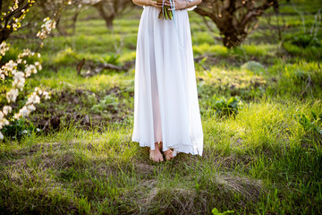 The girl in a white dress is standing with a bouquet of flowers in a meadow at sunset