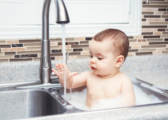 Portrait of cute Caucasian funny baby girl boy with dark black eyes sitting in big kitchen sink...