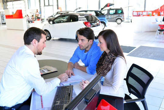 Vertragsabschluss im Autohandel - junges Paar kauft neuen PKW, Verkaufsgespräch und Vertragsunterzeichnung mit dem Händler // 
conclusion of contract in car dealership for sales