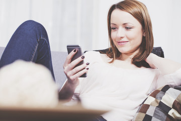 Beautiful smiling brown haired girl relaxing on sofa with smartphone in her hand