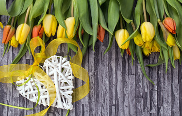 Wiosenny bukiet kwiatów z żółtych i czerwonych tulipanów oraz żonkili  na szarym tle z motywem muru i cegły z białym sercem ozdobionym żółtą wstążką zawiązaną na kokardkę