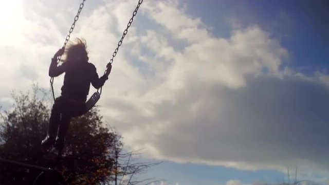 A mother pushes her son on a swing and runs underneath him, in slow motion, silhouette in the sky