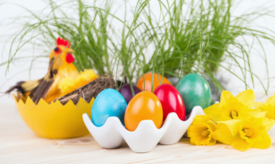 Pisanki, Wielkanocne dekoracje z kolorowymi jajami wielkanocnymi w ceramicznym stojaku, żonkilami i zielonymi roślinami w tle i kurczakiem  - obrazy, fototapety, plakaty
