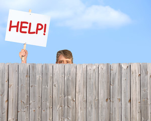 A man holding a HELP sign is peering over a fence desperate for help with his many problems in...