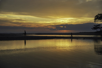 Fototapeta na wymiar silhouette image of two boys walking inline at the shore with beautiful sunrise sunset background. soft clouds and reflection on the water