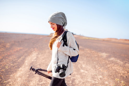 Young female photographer in sweater and hat walking with camera, backpack and tripod on the deserted landscape backround