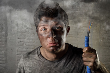 young man holding electrical cable smoking after electrical accident with dirty burnt face in funny sad expression