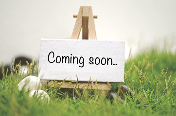 image concept white frame canvas on wooden tripod with word COMING SOON. Blurred and soft focus background with green grass and white stone - 104820221