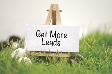 image concept white frame canvas on wooden tripod with word GET MORE LEADS. Blurred and soft focus background with green grass and white stone
