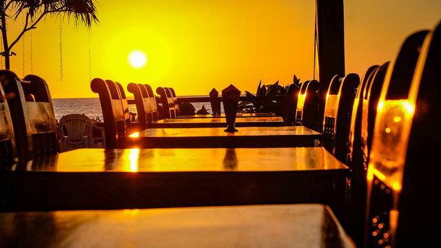 Empty beach restaurant with dark wood tables and dark wood chairs in front of a yellow sunset