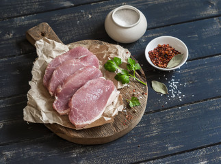 Raw pork chops and spices on a rustic wooden cutting board on a dark wooden background