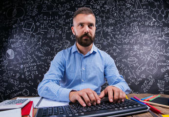 Happy teacher sitting at desk, school supplies, big blackboard
