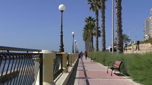 Malta island.People walking along Mediterranean Bugibba tropical resort promenade, bus passing.