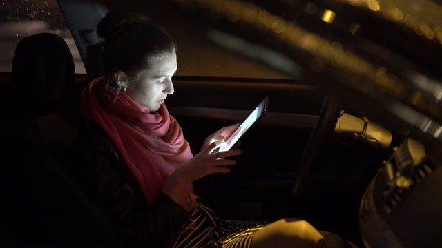 Pretty girl in a skirt sitting in car, smiling and using tablet at night, 4K