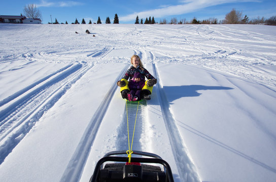 A teenage and a toddler girl being pulled behind a snowmobile on a sleigh in winter