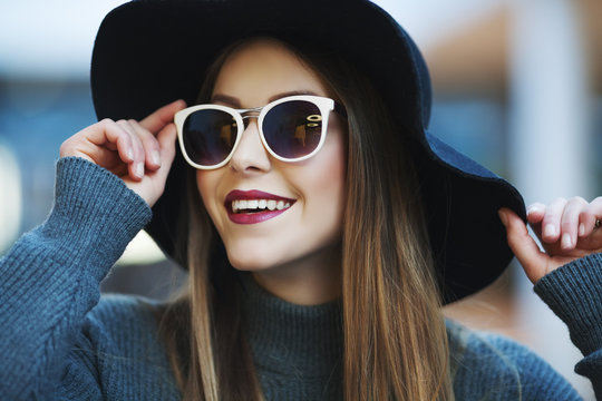 Close up portrait of a young beautiful smling woman with sunglasses and wide-brimmed black hat. Model looking at camera. Female fashion concept