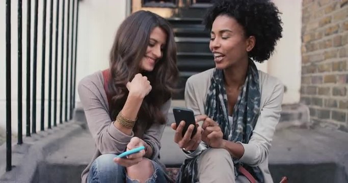 Two beautiful woman friends sitting on steps having fun using smart phone