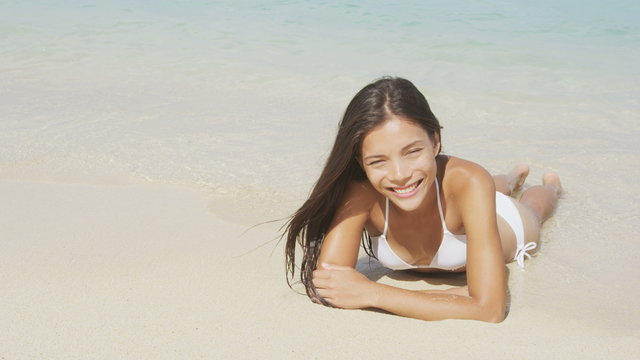 Beach bikini girl lying on beach sand in water in tropical paradise looking at camera. Sexy happy smiling woman in relaxing in the sun tanning. Beautiful multicultural Asian Caucasian model. 