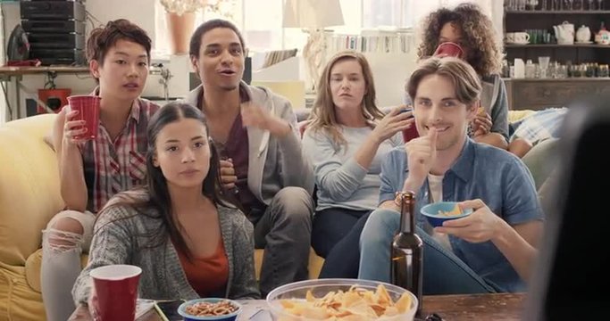 Diverse group of student friends arms raised celebrating goal watching sports event on TV together eating snacks  drinking beer 