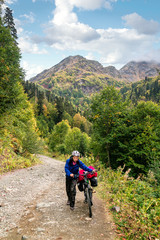 Fototapeta premium Young woman hiker and bicycle walking on countryside terrain. Tourist emerging from a lengthy uphill, mountains in the background