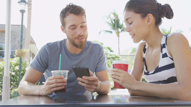 Smart phone Cafe couple looking at smartphone screen app laughing having fun on date drinking coffee in summer. Young man using talking with Asian woman sitting outdoors. Friends in late 20s.