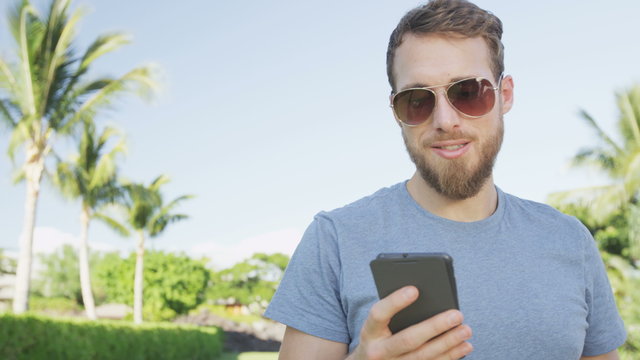 Man sms texting using app on smart phone outdoors in summer. Handsome young casual man using smartphone smiling happy wearing sunglasses. Urban male hipster. RED EPIC 90 FPS.