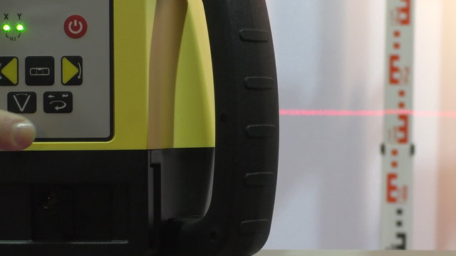 Close up of human finger and buttons on rotary laser level and grade rod or staff and red line in the background